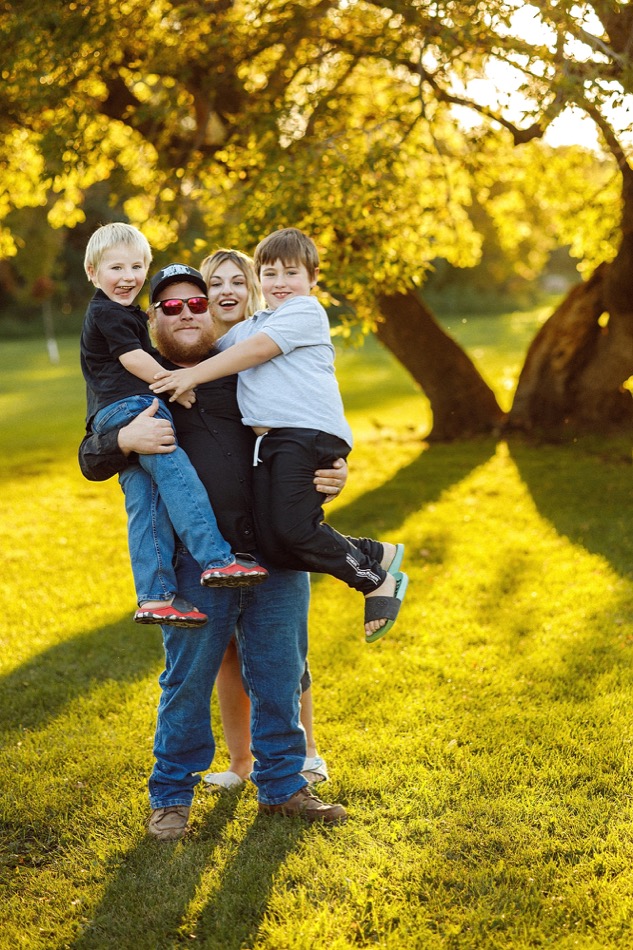 Father holding two smiling boys with their mother behind them in a sunlit park