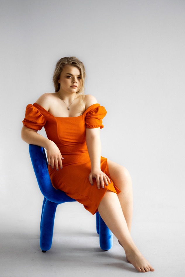 Woman in an orange dress seated on a blue chair in a studio