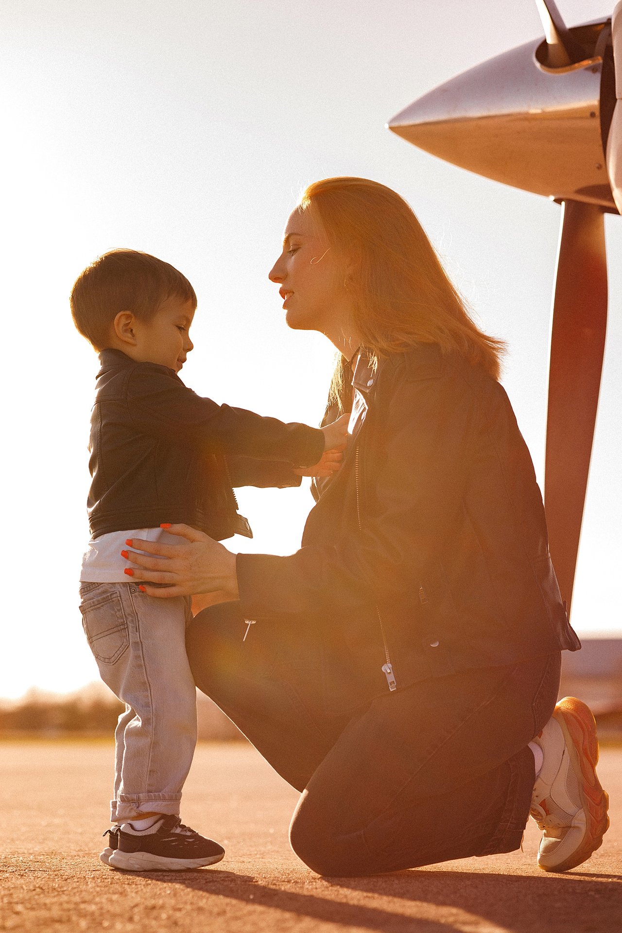 Alina crouching face to face with David beside the propeller of a small aircraft.