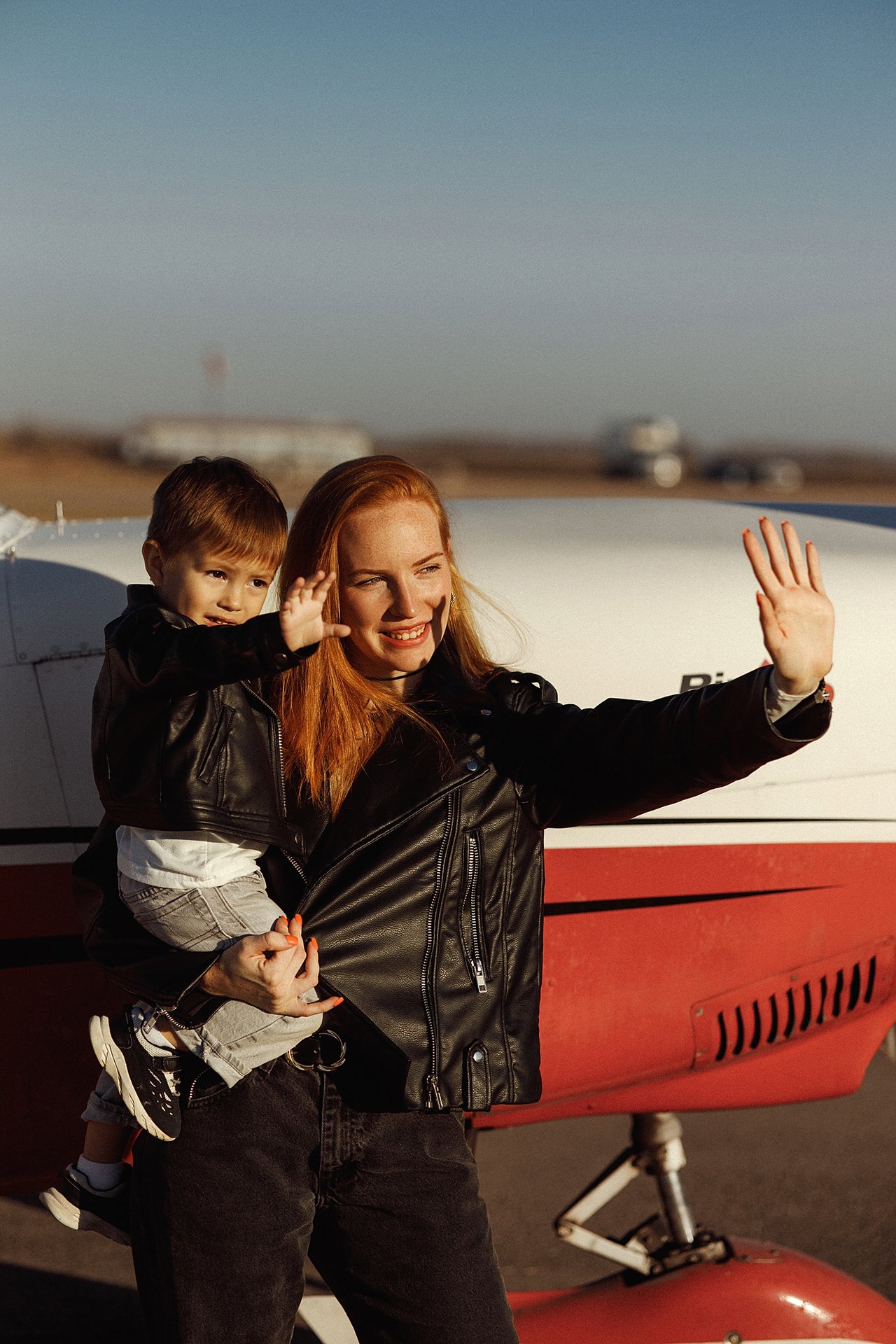 Alina holding David as they both wave beside a red and white aircraft.