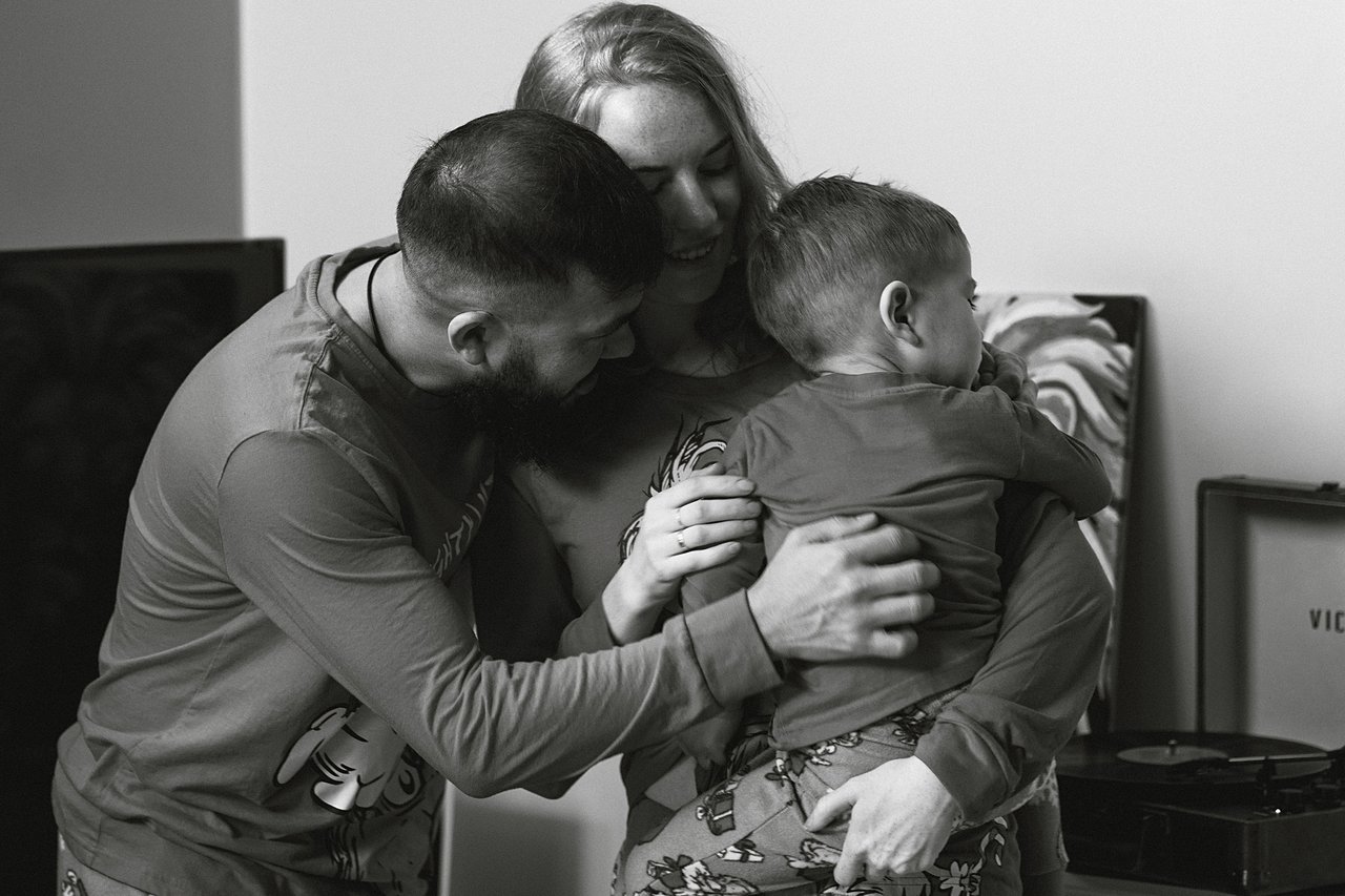 Black and white photograph of Alina, Nurali and David sharing a close family embrace at home.