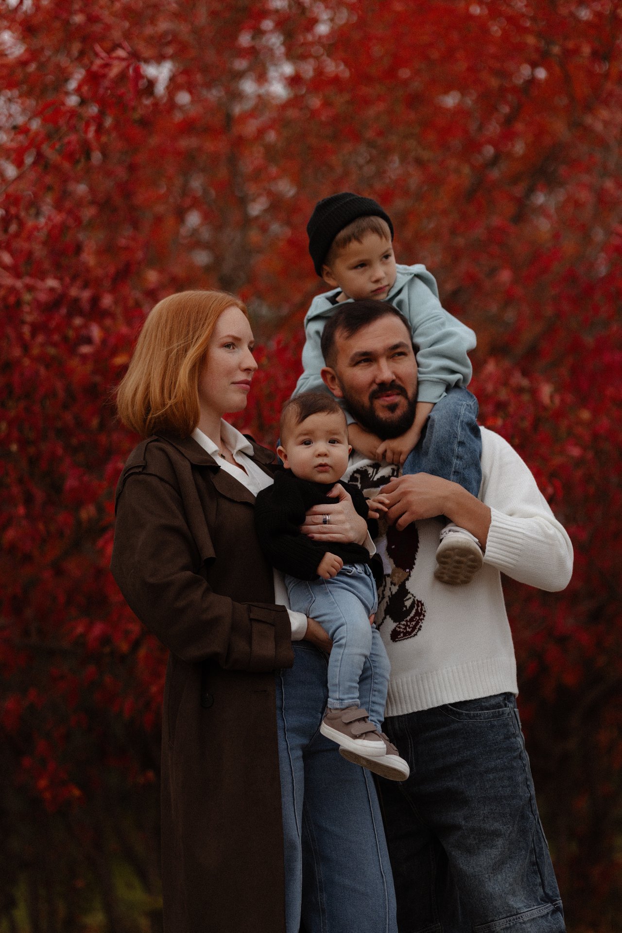 Alina and Nurali with both children, with David on Nurali’s shoulders against red autumn leaves.